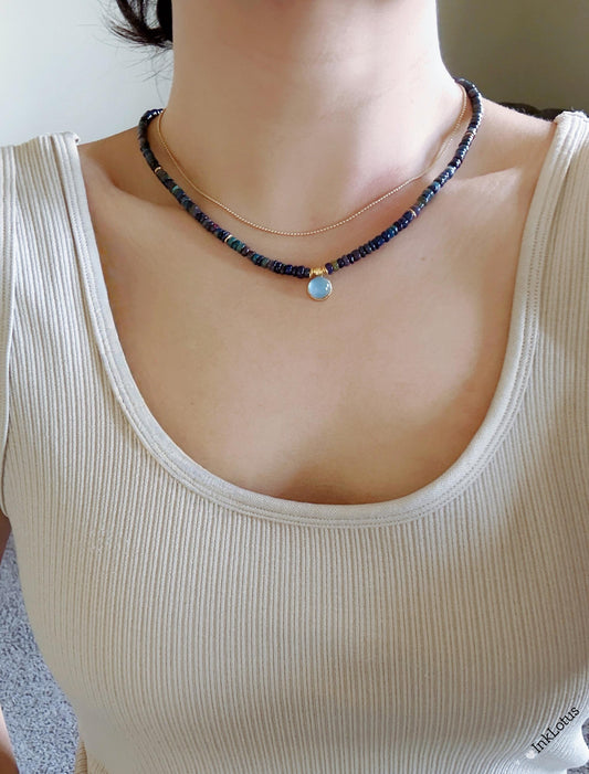 May include: Two necklaces. The top necklace is a delicate gold chain. The bottom necklace features small, dark blue beads and a small, round, light blue pendant with a gold border. The necklaces are worn on a person wearing a beige tank top.